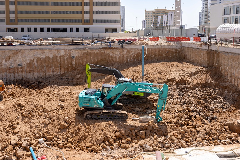 Cove Edition I By Imtiaz 1 A construction site featuring a Kobelco excavator digging in a large dirt pit near a building structure.